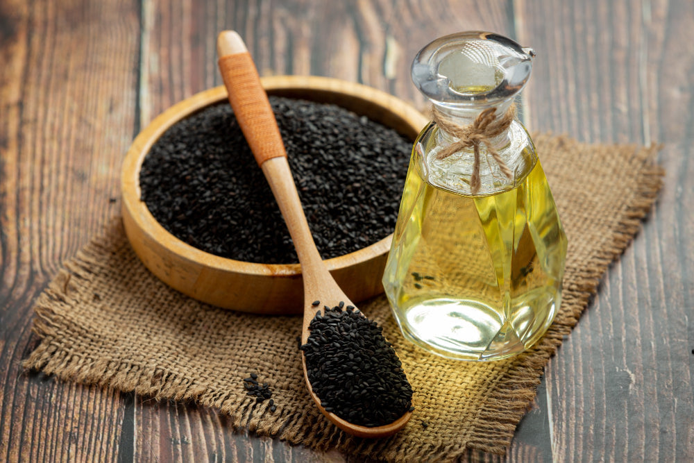 Black sesame seeds scattered beside a small glass dish of dark pressed oil on a dark textured background.