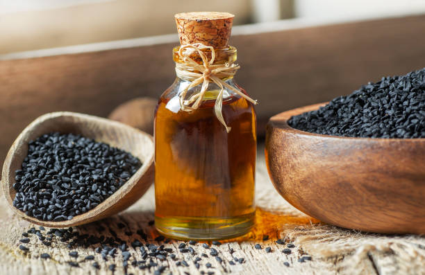  Black seed oil in a glass bottle with cork stopper surrounded by nigella sativa seeds in wooden bowls.